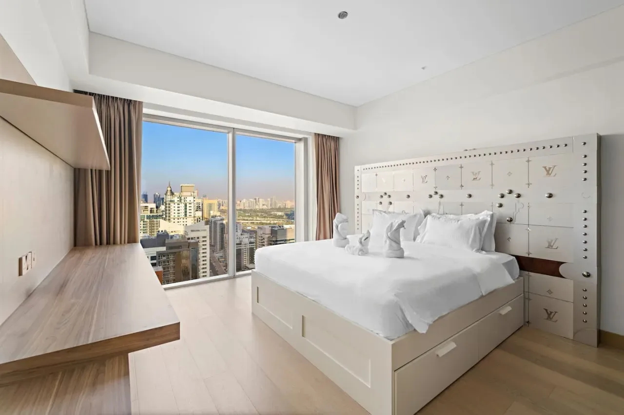 A bright bedroom with a neatly made bed beside a large window overlooking the city.
