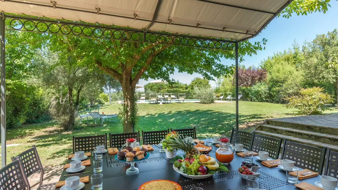 Una mesa de comedor al aire libre bajo techo da a un jardín con árboles y césped.