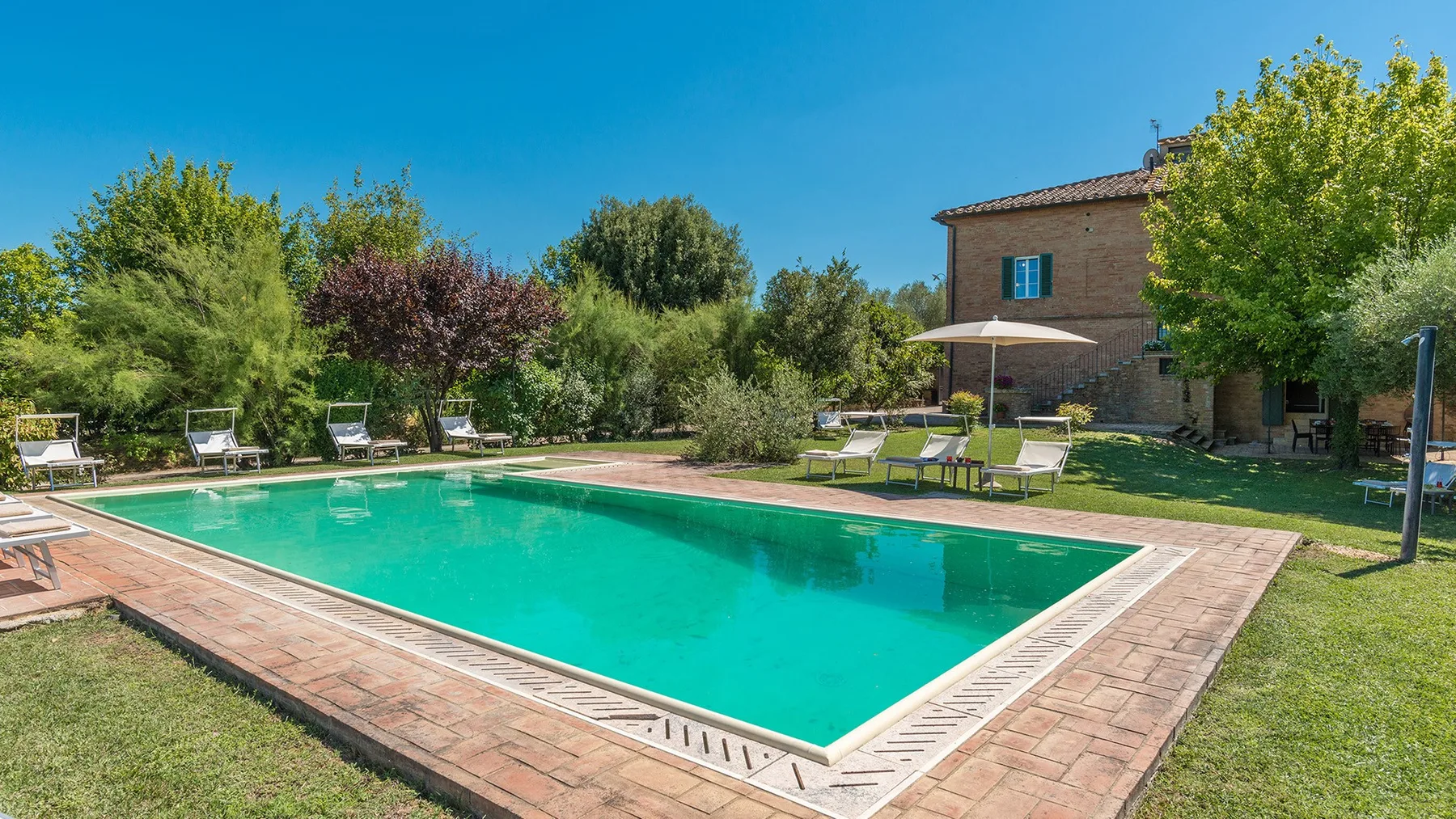 Una piscina al aire libre está junto a un césped, árboles y una casa.