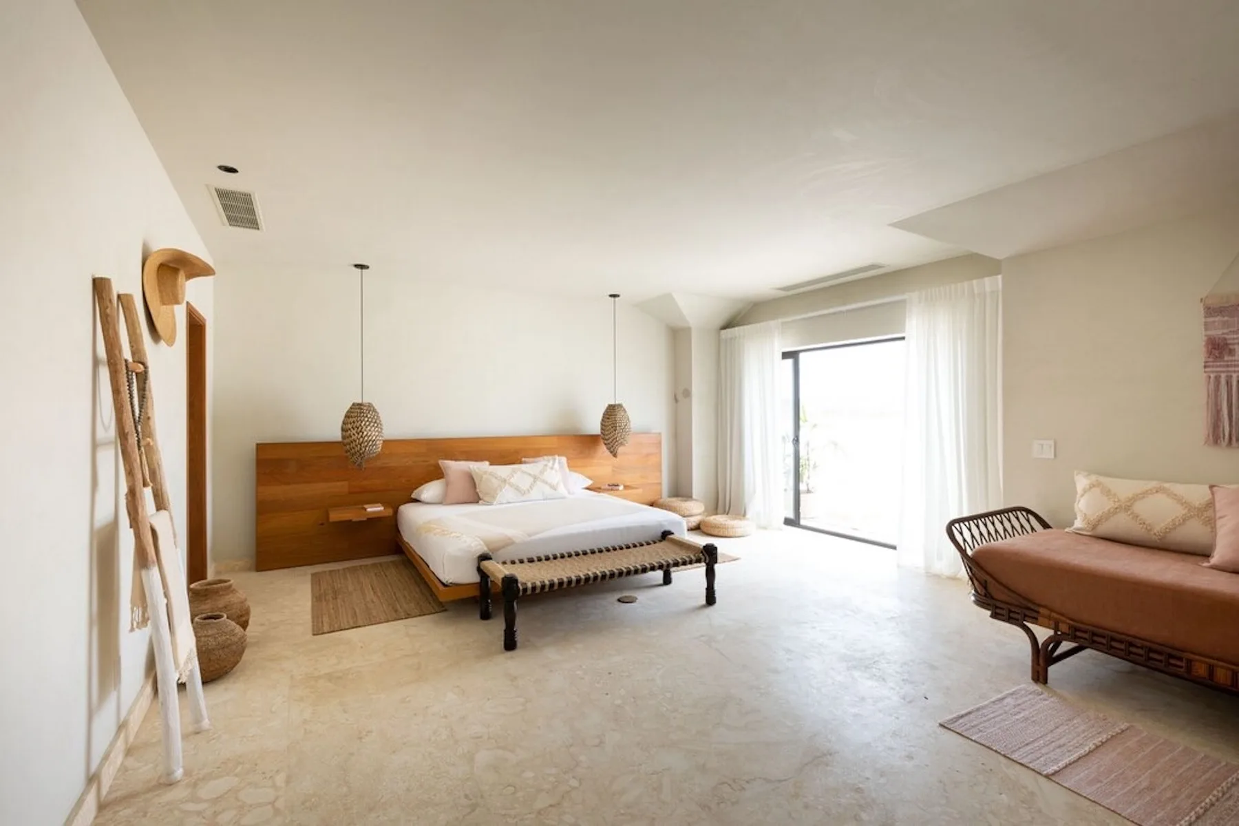 A bright bedroom with a bed, woven bench, and seating beside large windows.