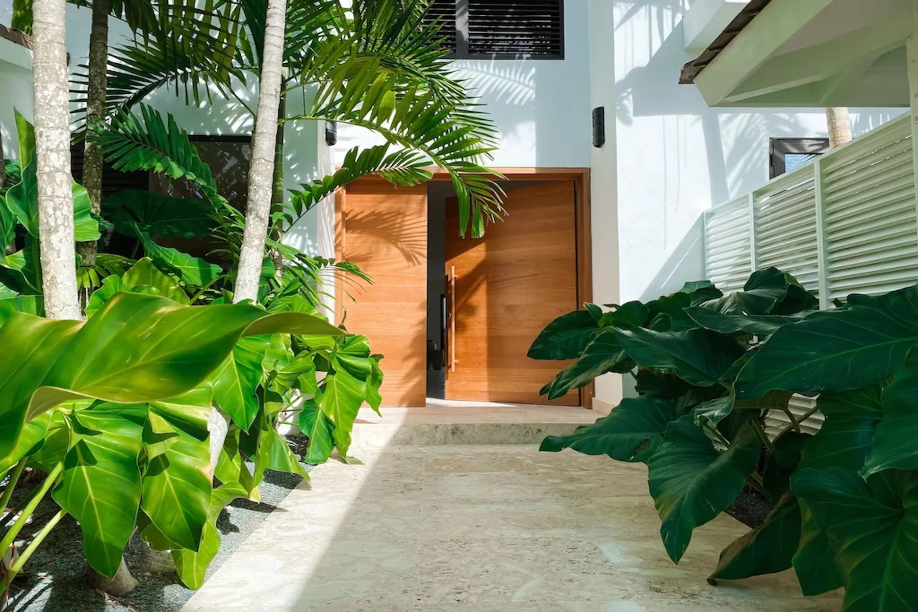 A leafy entrance walkway leads to a wooden doorway between tropical plants.