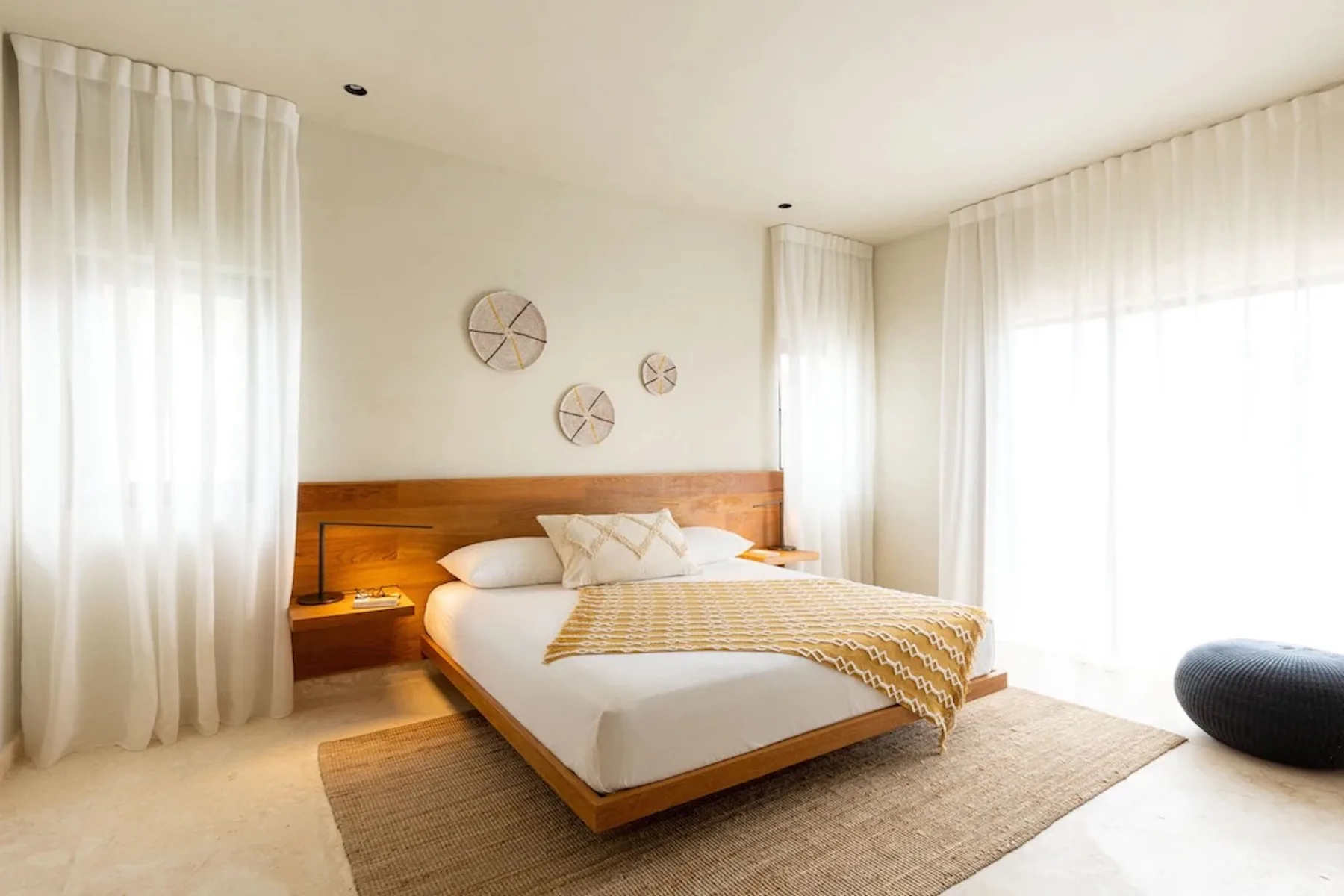 A bright bedroom with a low wooden bed and sheer curtains.