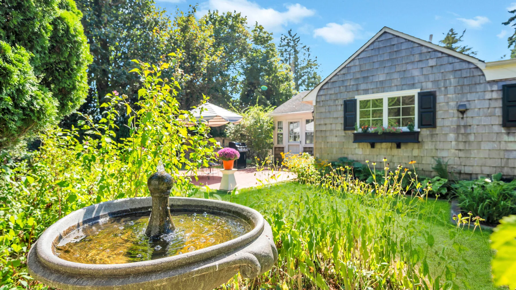 Una fuente de jardín está junto a una casa de tejas, rodeada de abundante vegetación.