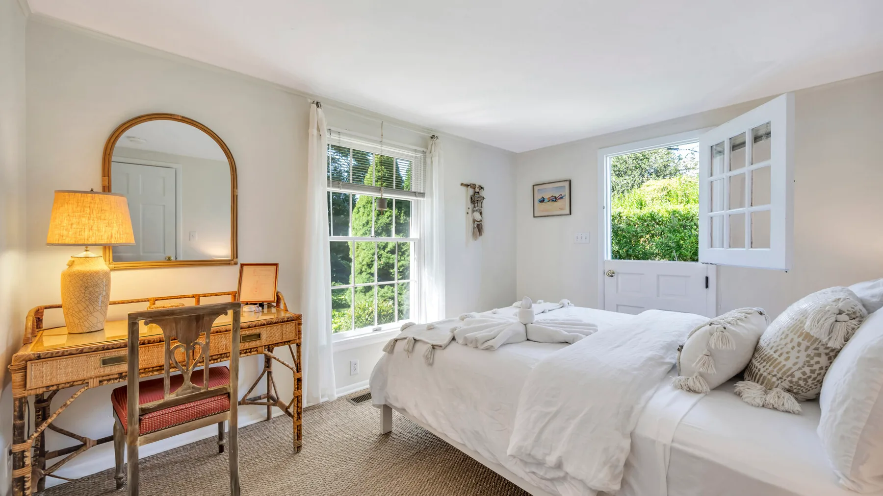 Un dormitorio luminoso con una cama blanca, un escritorio y ventanas grandes.
