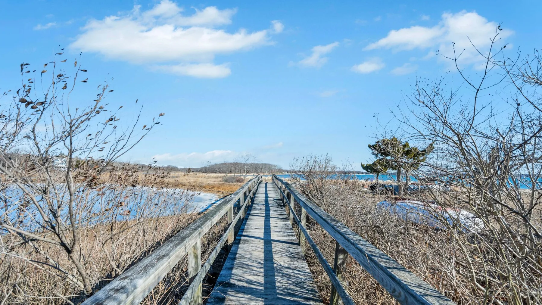 A wooden boardwalk crosses a marsh toward the water under a blue sky.