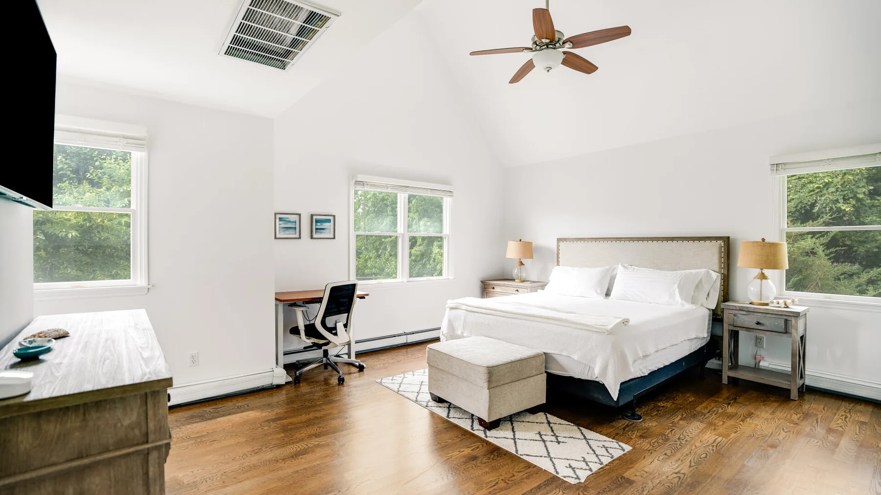 A bright bedroom with a bed, desk, and windows overlooking greenery.