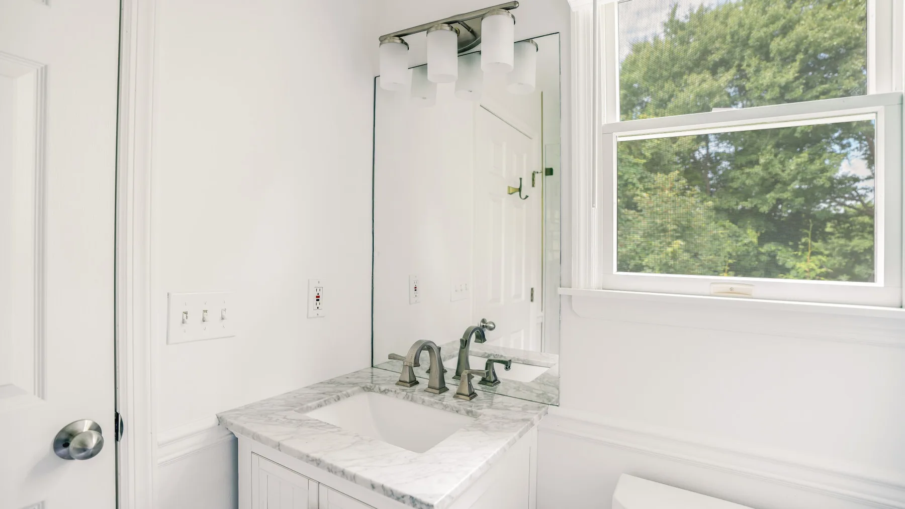 A bright bathroom vanity with a mirror sits beside a window overlooking trees.