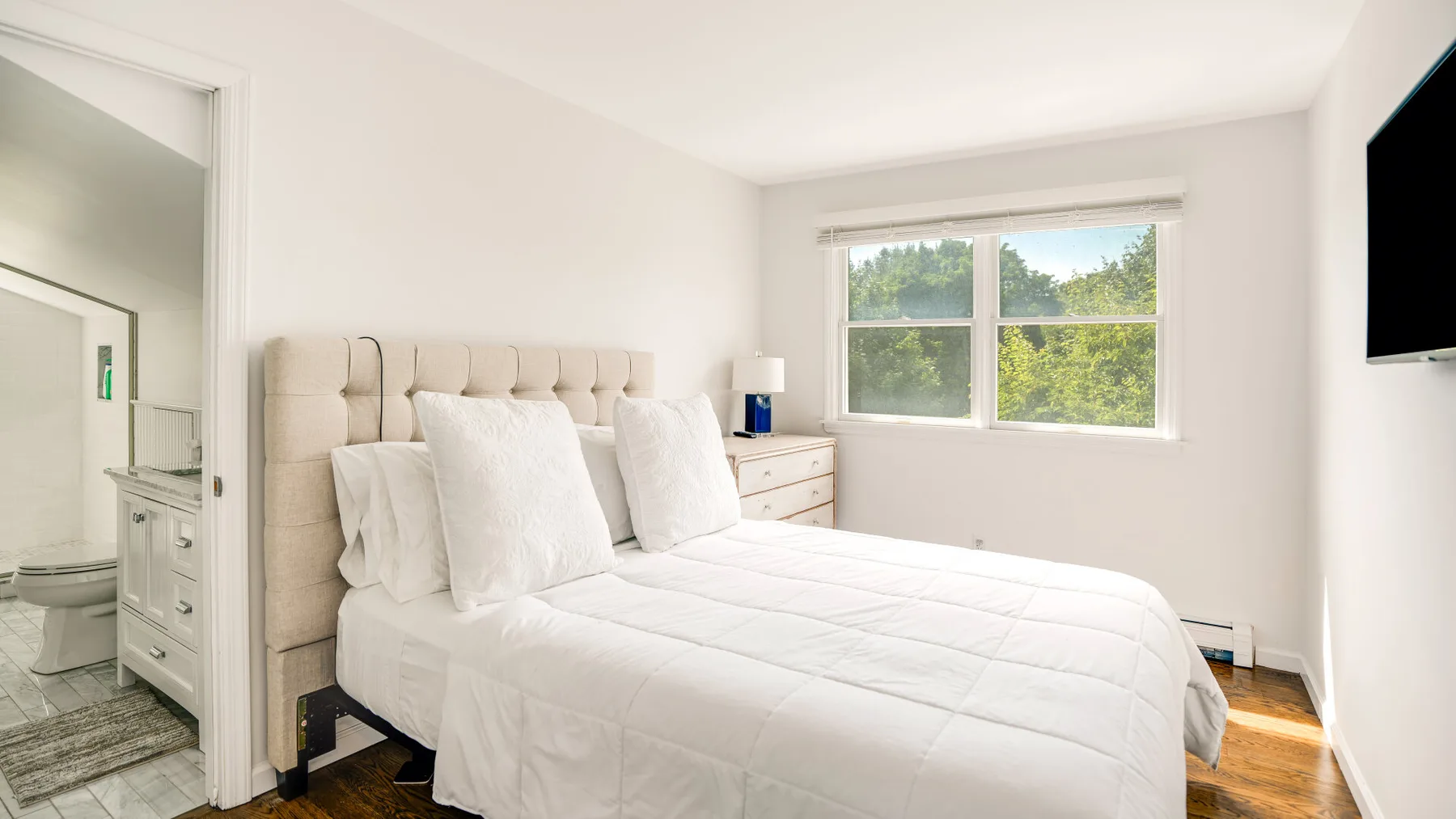 A bright bedroom with a white bed beside a window.