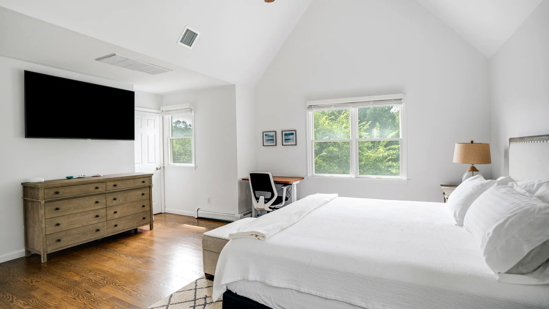 A bright bedroom with a large bed, wooden dresser, and windows overlooking greenery.