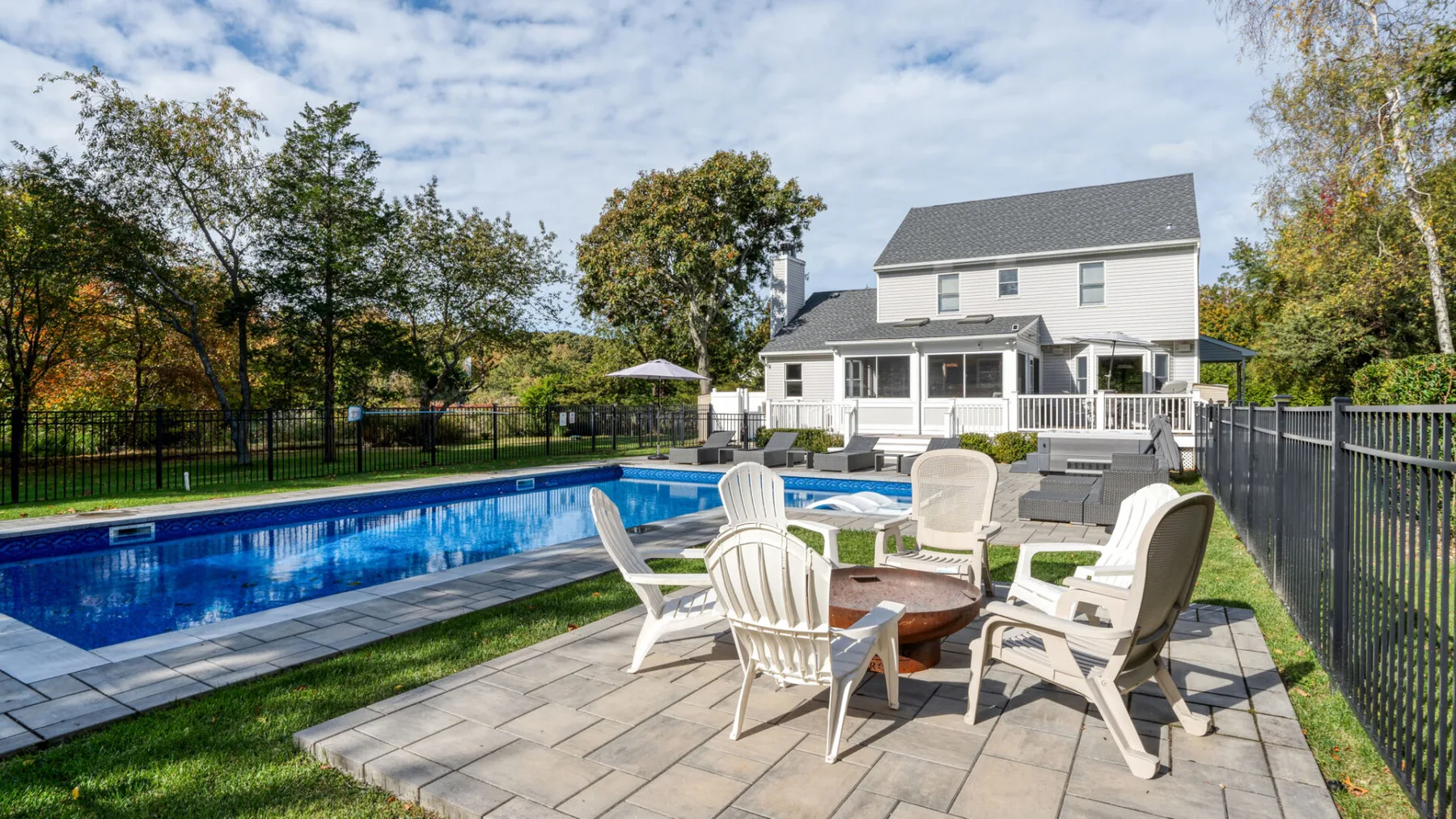 A backyard pool and patio seating sit beside a large white house.