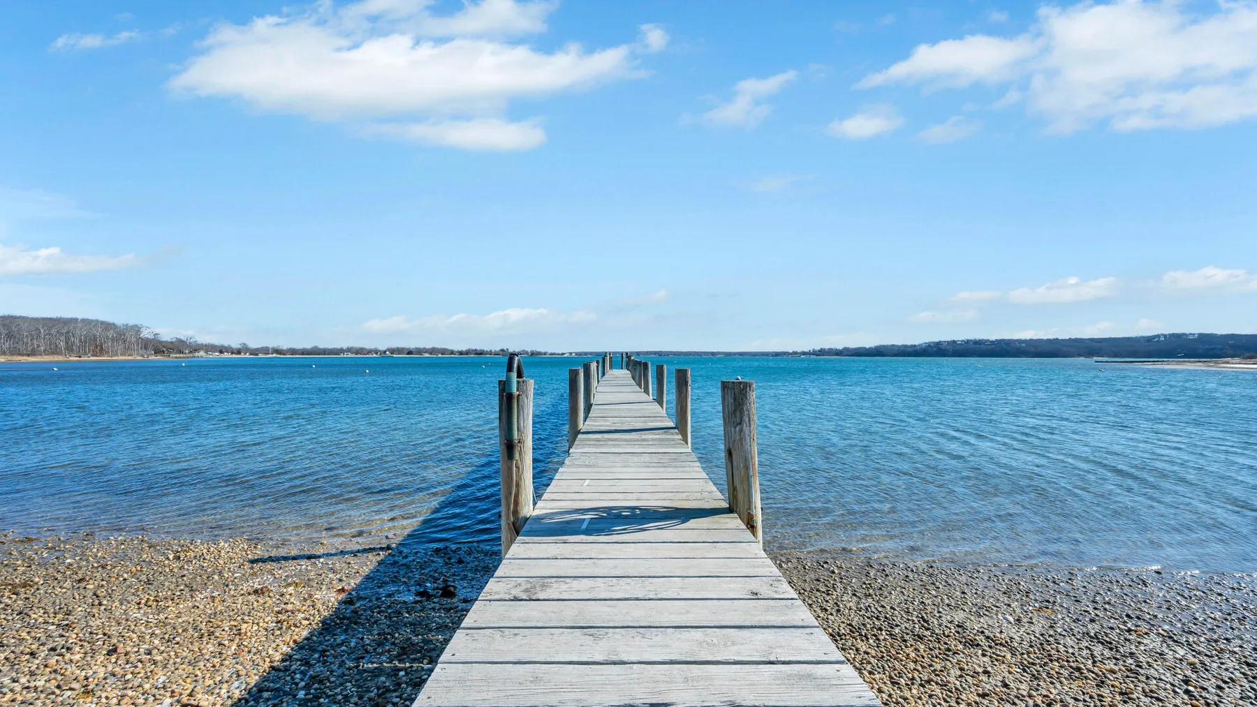 A wooden pier extends across a pebbled shore into a calm blue lake.