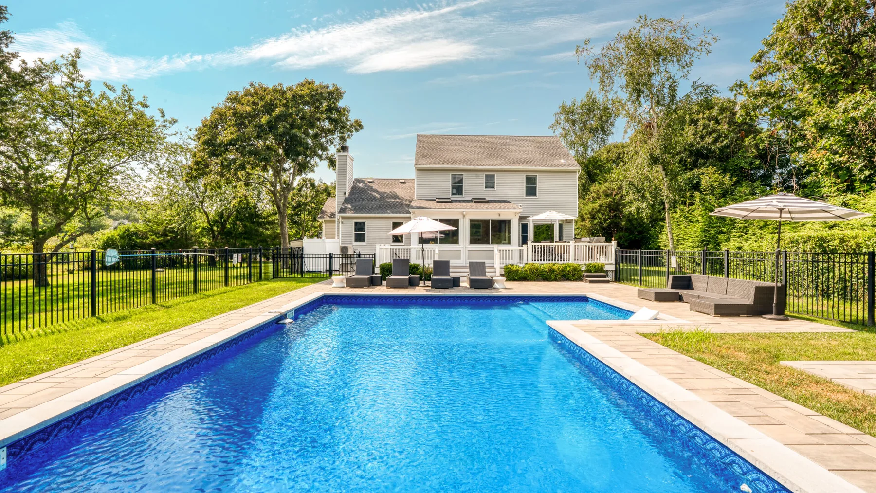 Outdoor pool area beside a house, with lounge seating and a fenced lawn.