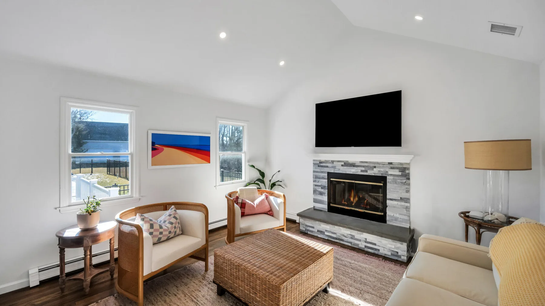 A bright living room with seating, a woven coffee table, and a fireplace beneath a television.