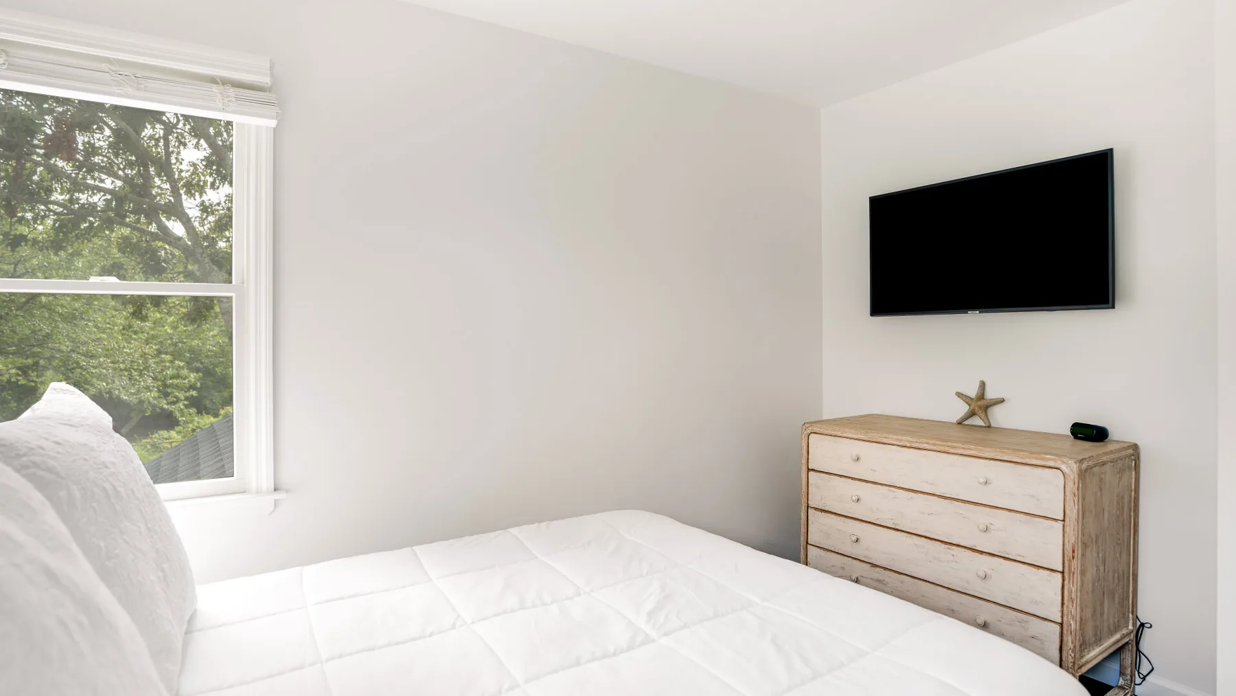 A bright bedroom with a white bed and a dresser beneath a wall-mounted television.