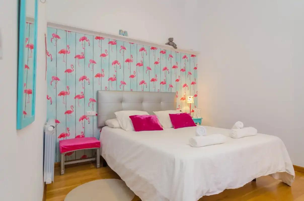 A bedroom with a white bed, pink pillows, and flamingo-patterned wallpaper.