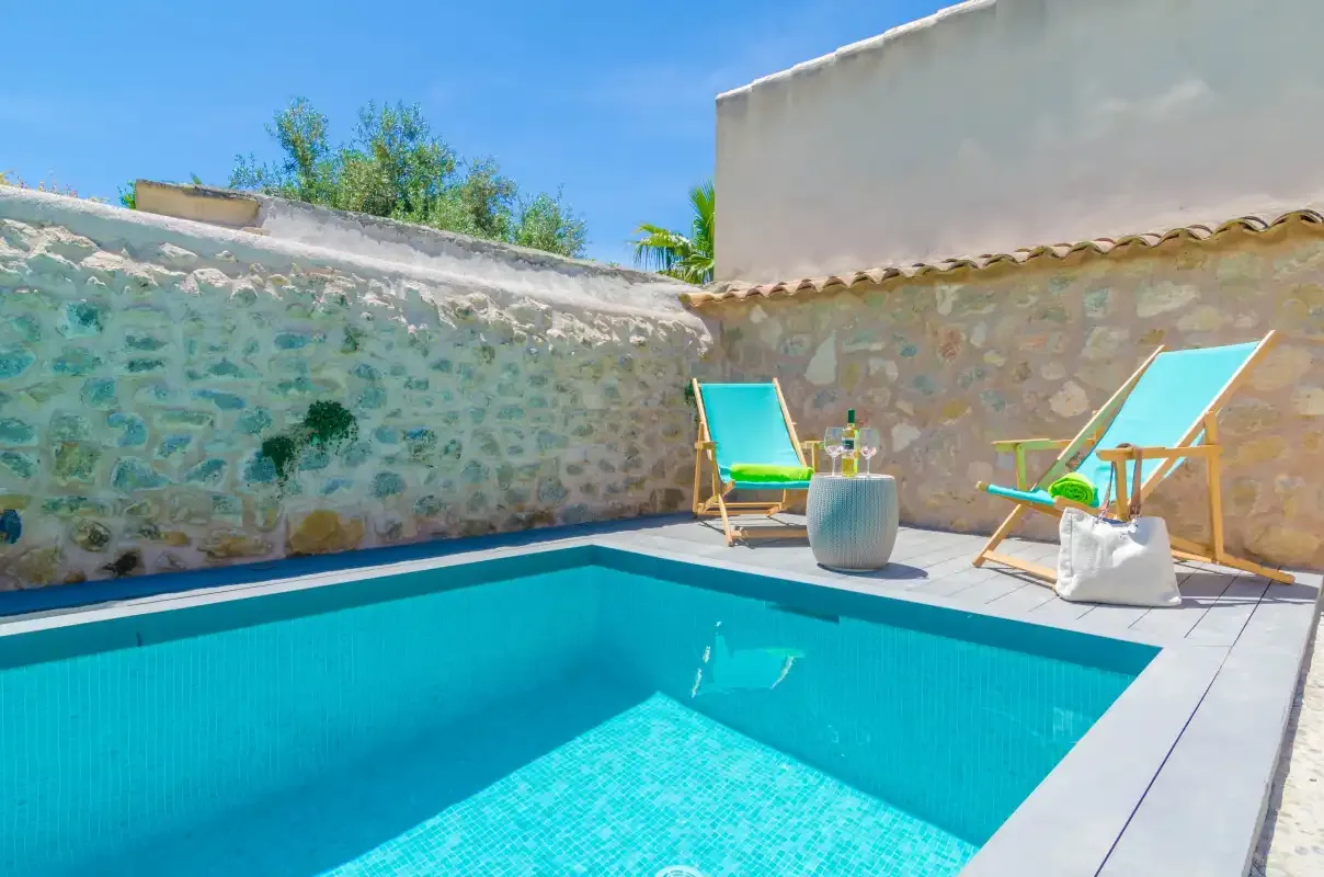 A swimming pool beside a stone courtyard wall and two lounge chairs.