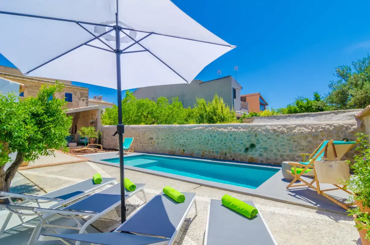 An outdoor swimming pool beside a stone wall, with loungers under a large umbrella.