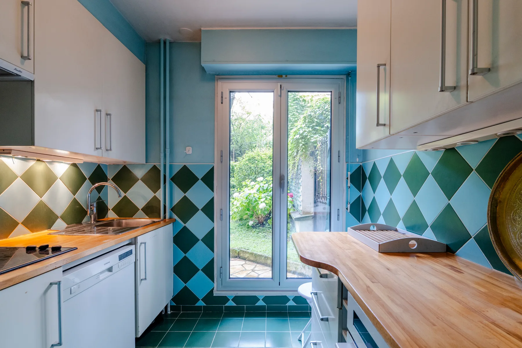 A narrow kitchen with wooden countertops, turquoise cabinets, and glass doors opening to greenery.