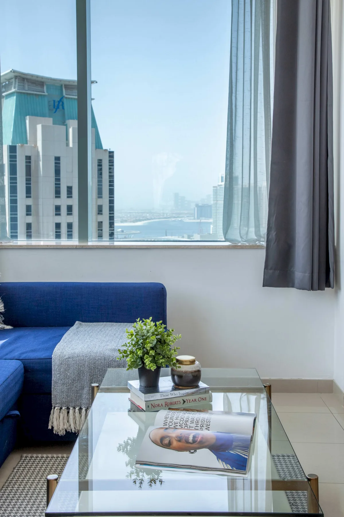 Una sala de estar con asientos azules y una mesa de centro de vidrio junto a una ventana grande.