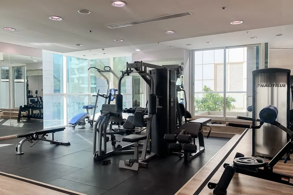 Una sala de ejercicios con máquinas y un banco de pesas junto a grandes ventanas.