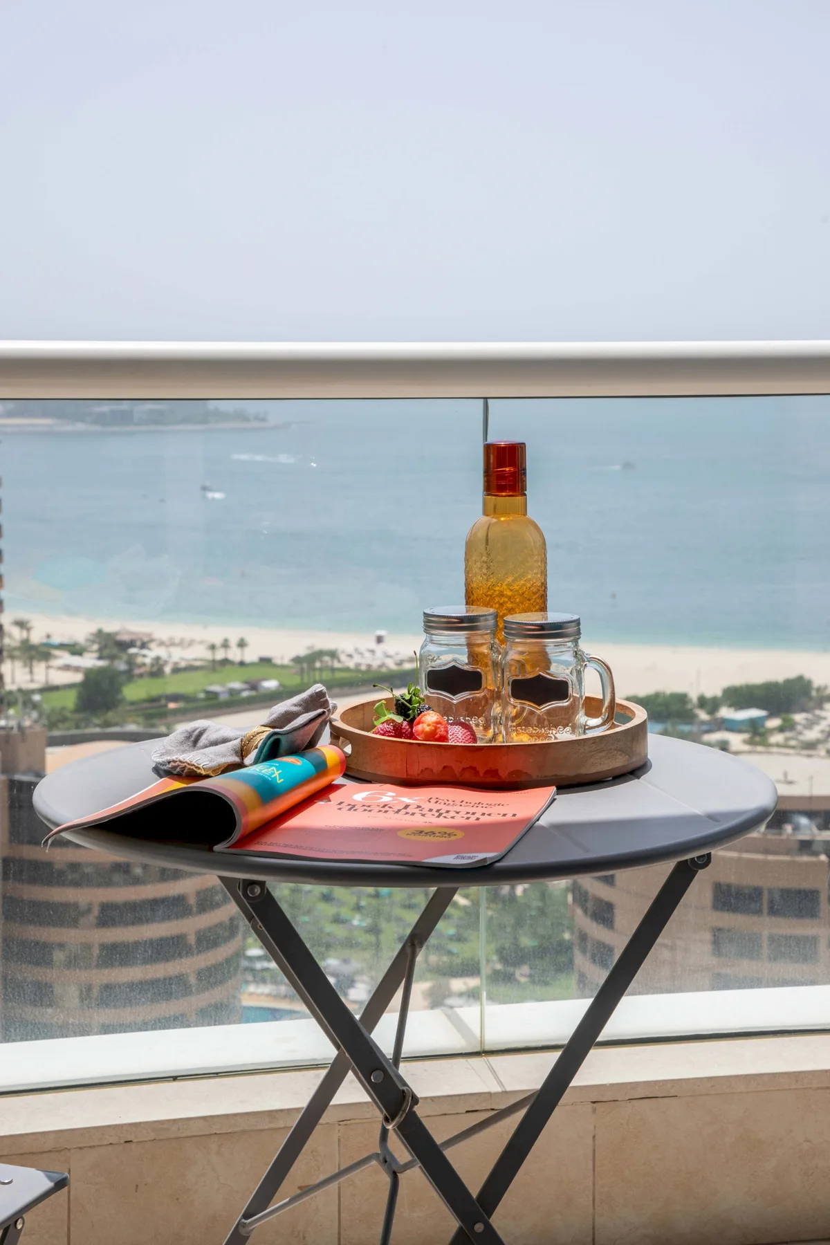 Una mesa pequeña con bebidas y material de lectura está frente a una vista al mar desde el balcón.