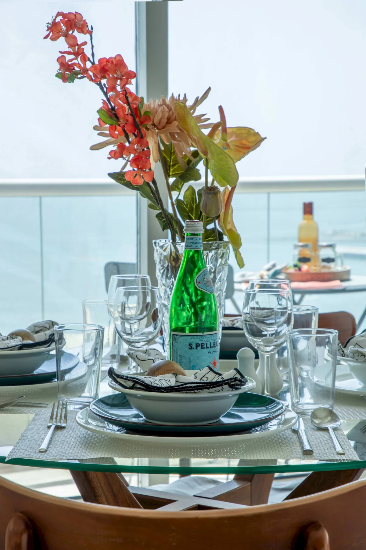 Una mesa de comedor está puesta con vasos, platos y un jarrón de flores junto a una ventana.