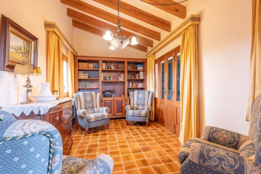 A sitting room with upholstered seating, a bookcase, and warm-toned tile flooring.