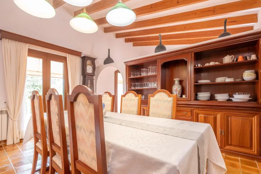 A dining room with a long table and wooden chairs beneath exposed ceiling beams.