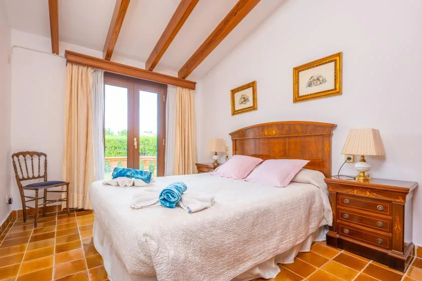 A bedroom with a large bed, wooden furnishings, and glass doors opening to greenery.