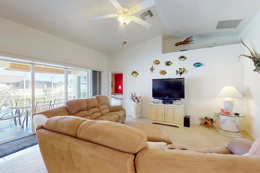A bright living room with tan seating, a television, and glass doors opening to a patio.