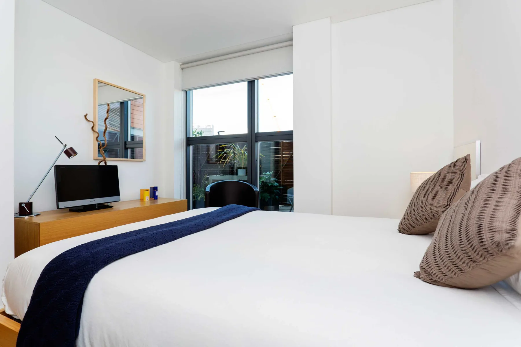 A bright bedroom with a neatly made bed and a desk beside a large window.