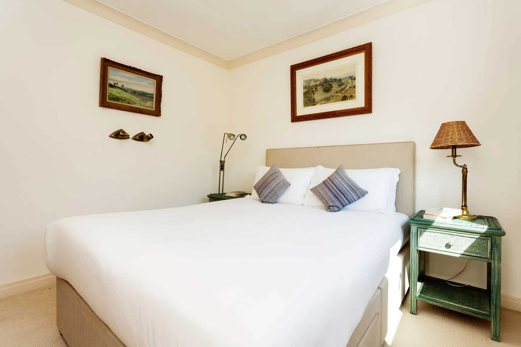 A bright bedroom with a neatly made bed and framed landscape artwork.