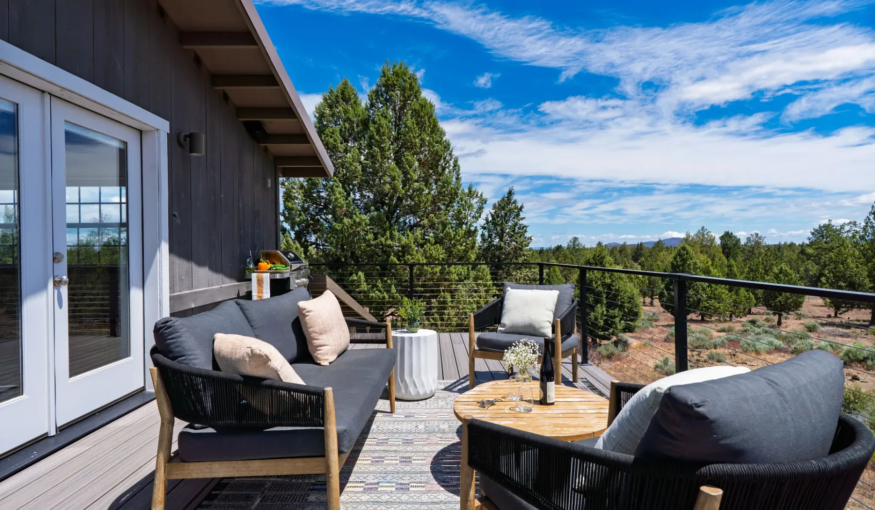 Un patio exterior tiene asientos y una mesa baja con vistas a un paisaje arbolado.