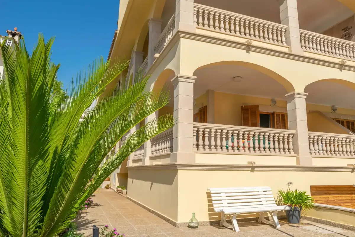 A cream-colored building with balconies stands beside a palm-lined paved walkway.