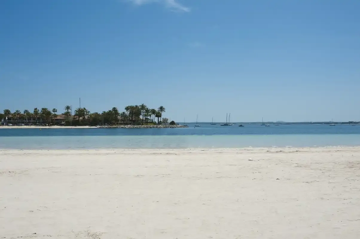 A sandy beach borders calm blue water, with boats visible offshore.