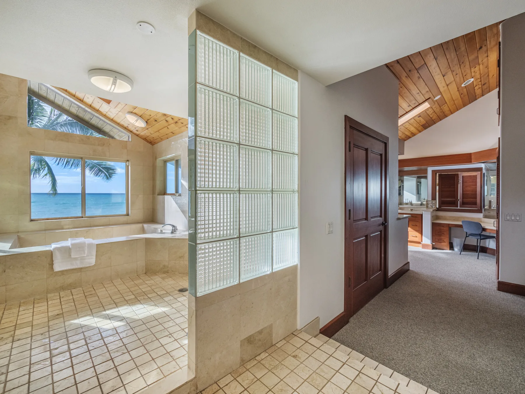 Un baño con azulejos, bañera y separador de bloques de vidrio junto a una habitación con techo de madera.