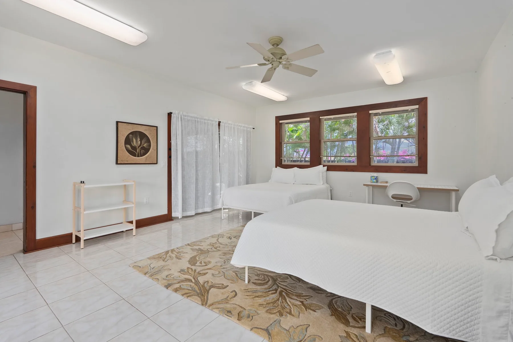 Un dormitorio luminoso con camas blancas, ventanas con cortinas y un escritorio pequeño.