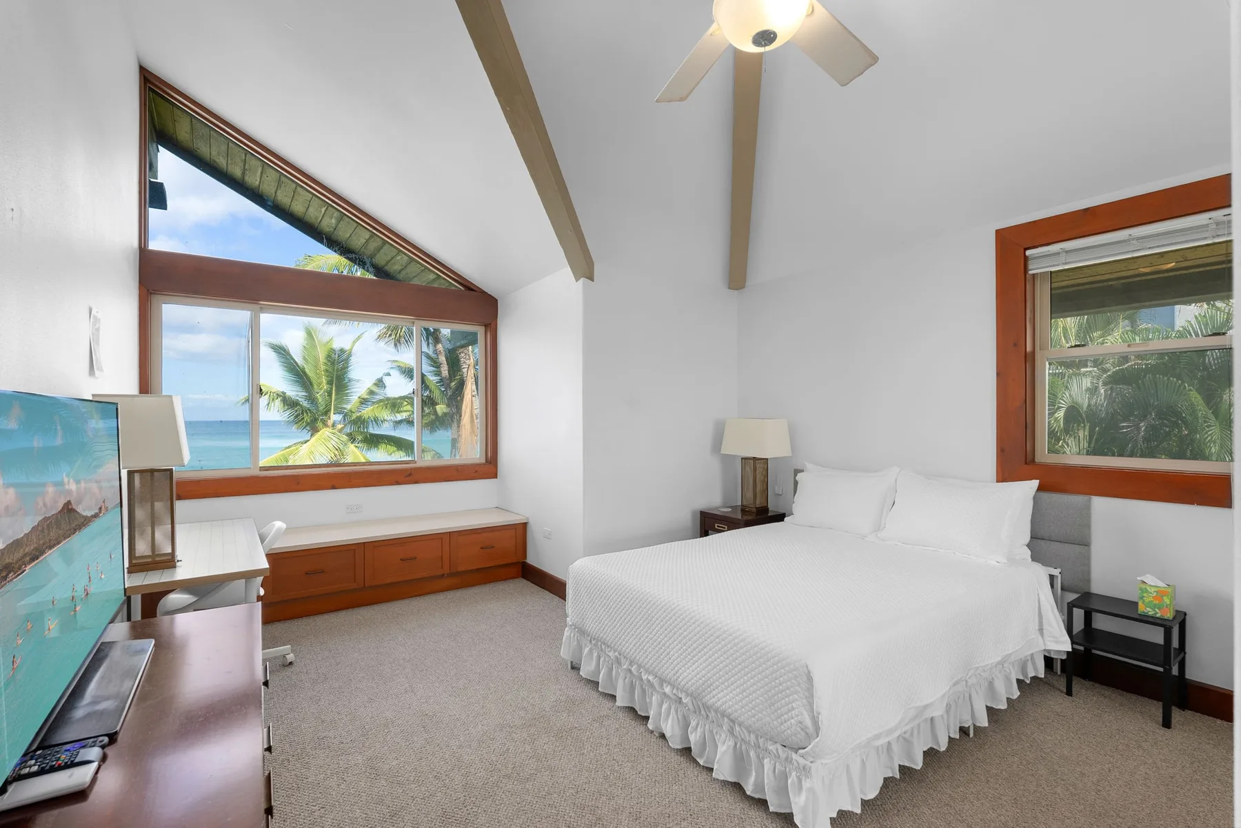 Un dormitorio luminoso tiene una cama blanca, un ventilador de techo y grandes ventanas con vista a palmeras.
