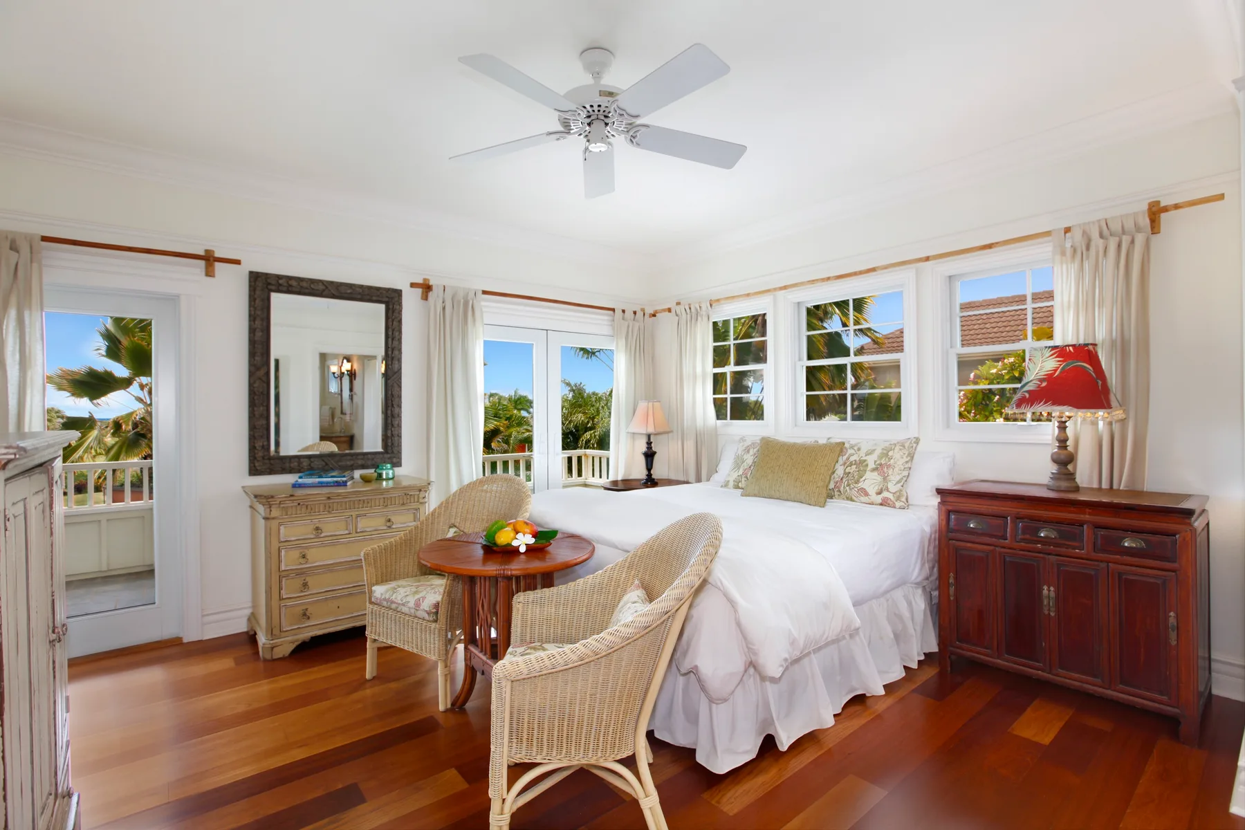 A bright bedroom with a bed, wicker seating, and windows overlooking greenery.