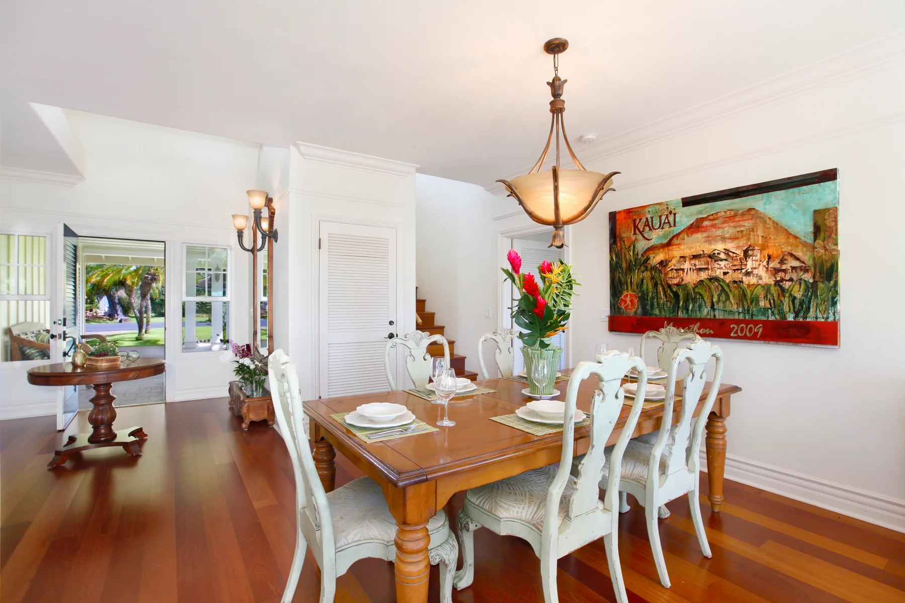 A dining room with a wooden table, white seating, and a colorful wall hanging.
