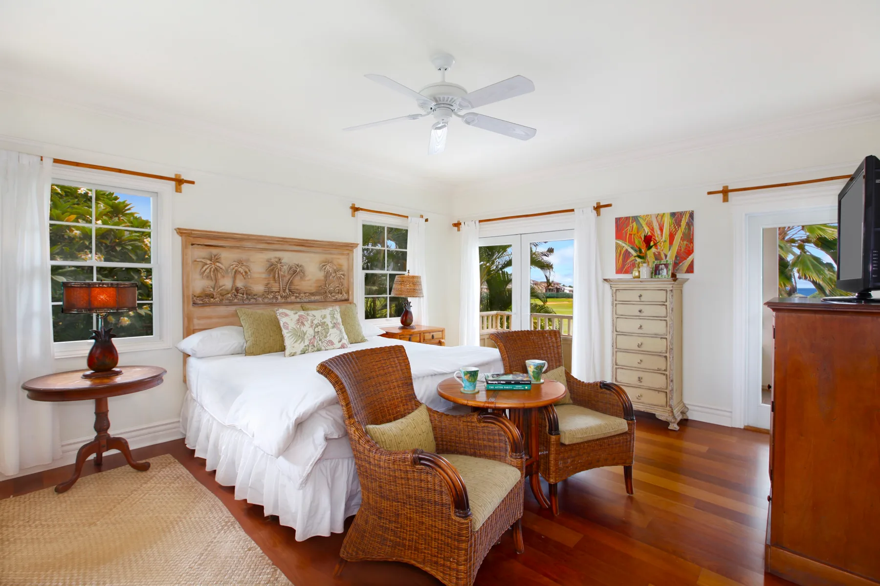A bright bedroom with a large bed, wicker seating, and windows overlooking greenery.