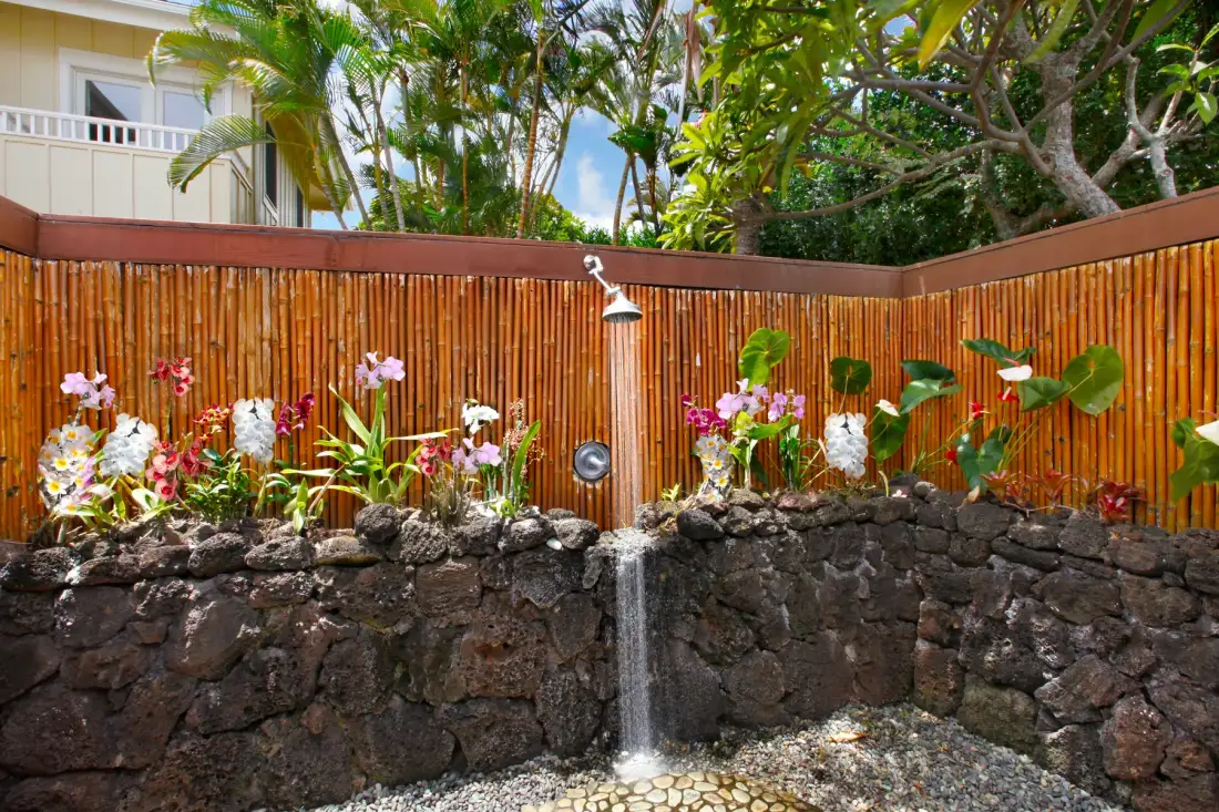 An outdoor shower stands beside a stone wall with tropical plants and flowers.