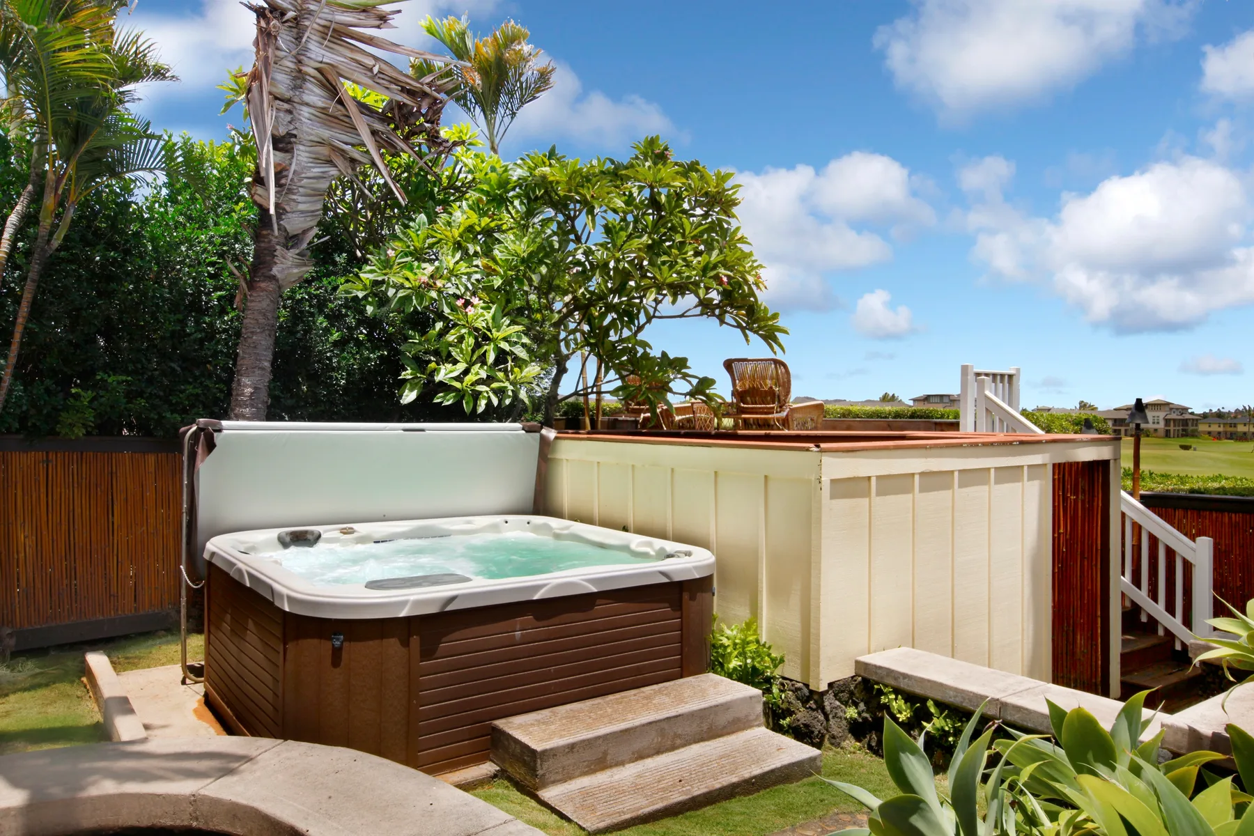 An outdoor hot tub sits beside a fenced deck under a blue sky.