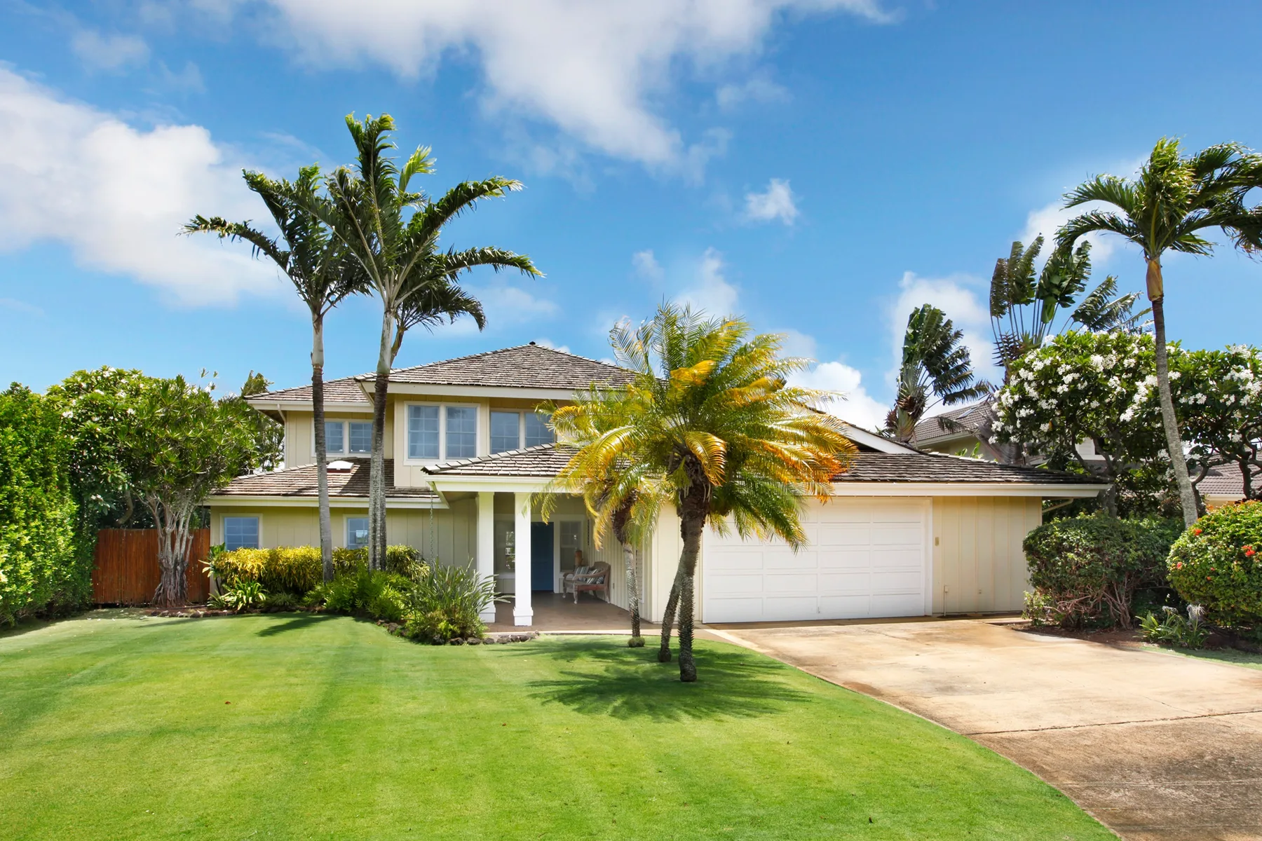 A two-story house stands behind a green lawn, with palm trees and a driveway.