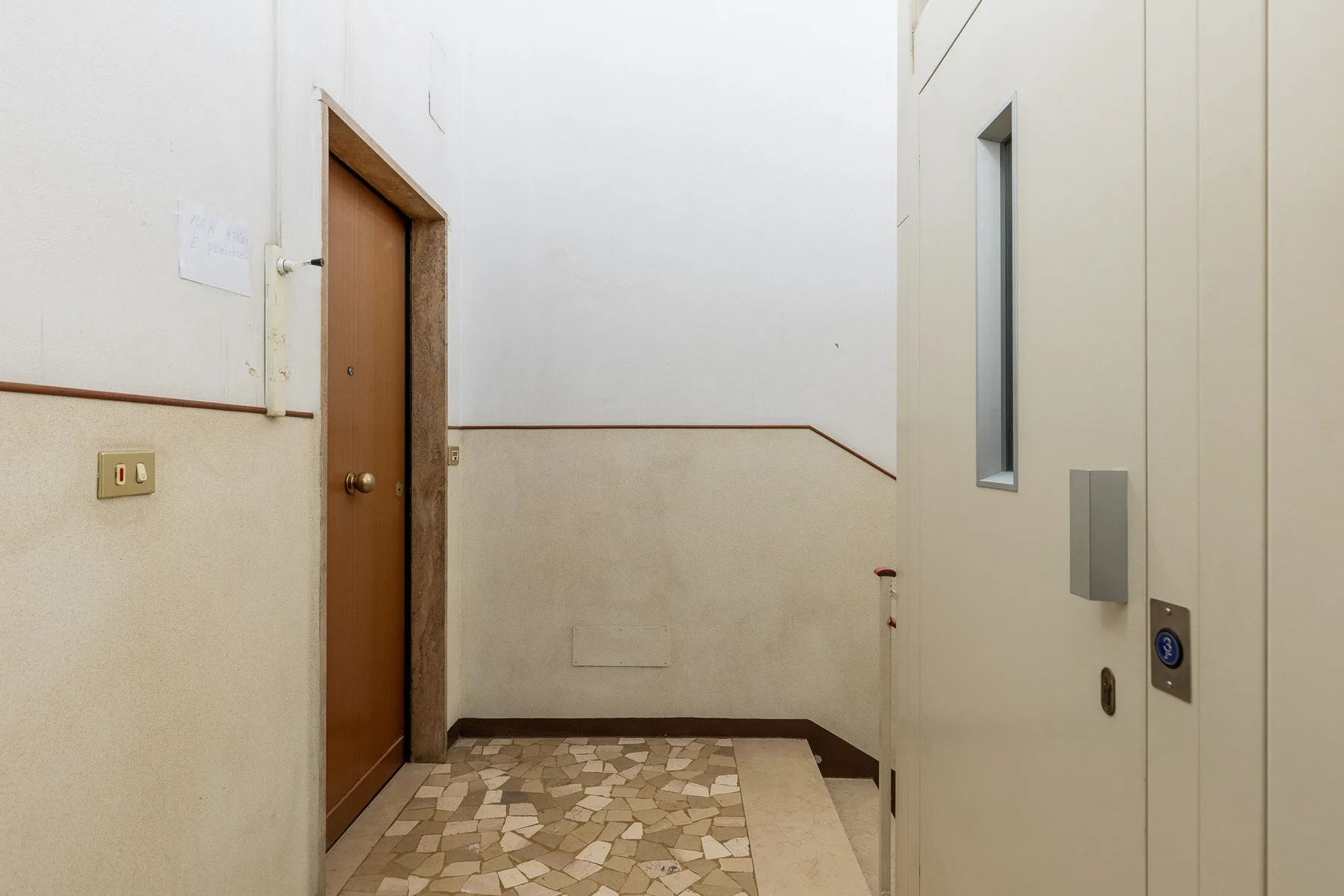 Un pasillo interior luminoso tiene una puerta de madera y un ascensor.