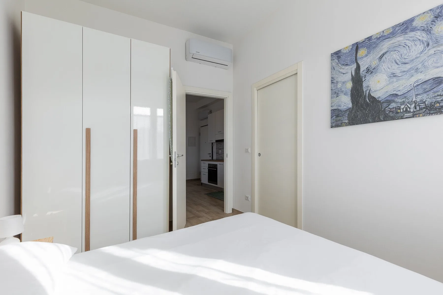 Un dormitorio luminoso con una cama blanca, armario empotrado y una obra enmarcada.