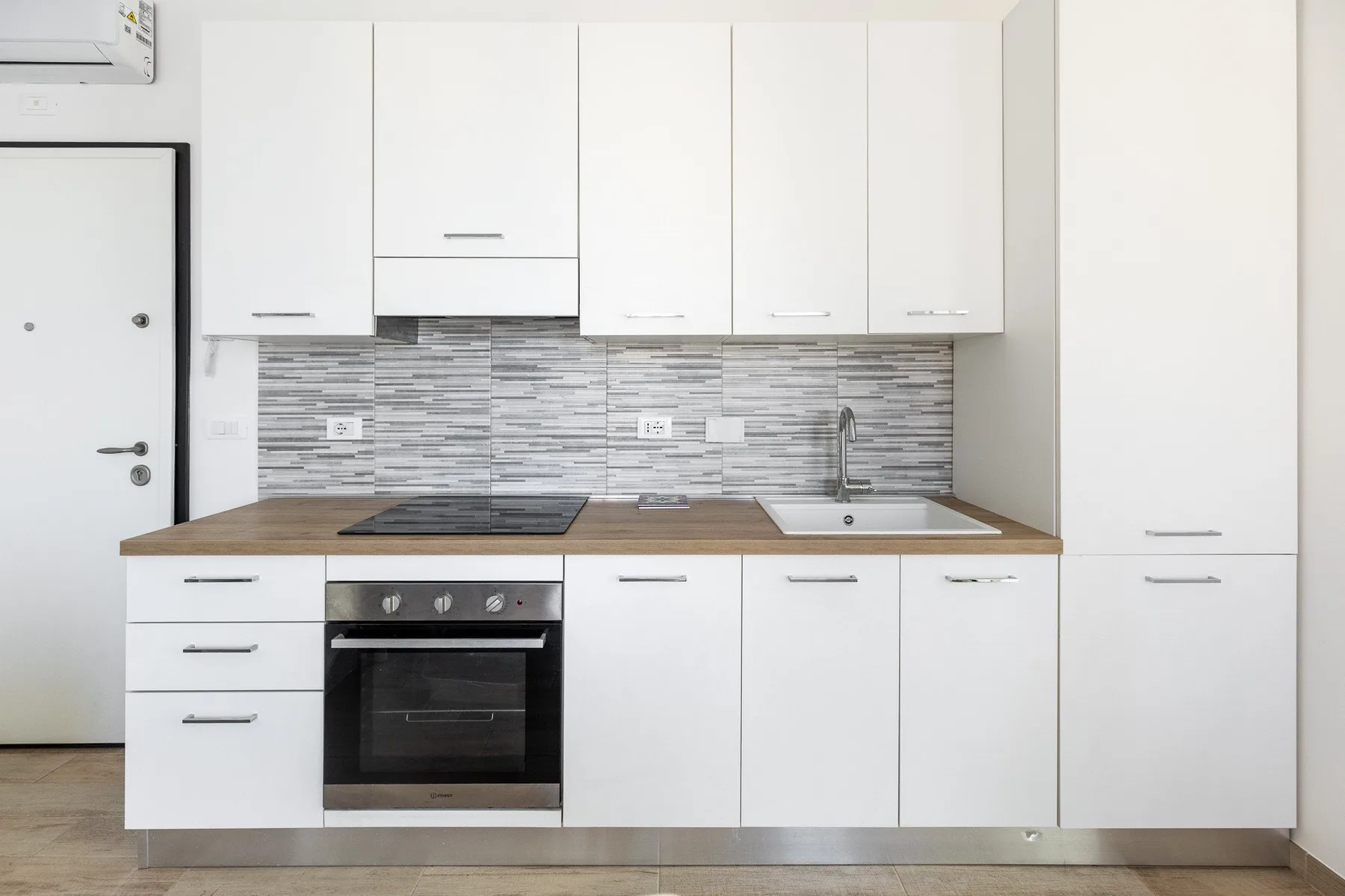 Una cocina compacta blanca con placa de cocción, horno, fregadero y armarios superiores.