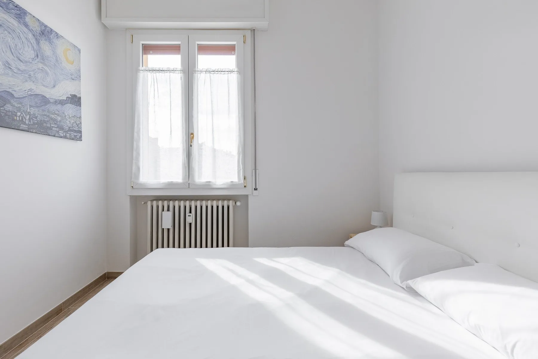 Un dormitorio luminoso con una cama blanca bajo una ventana con cortinas.