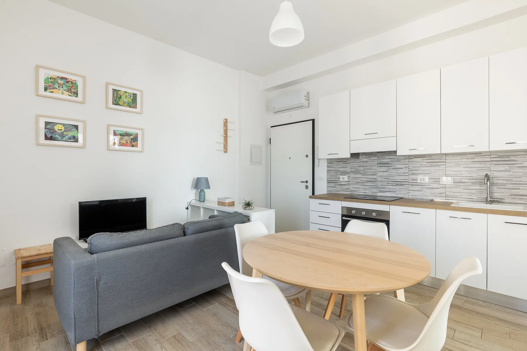 Una sala de estar y comedor con sofá gris, mesa redonda y gabinetes de cocina blancos.