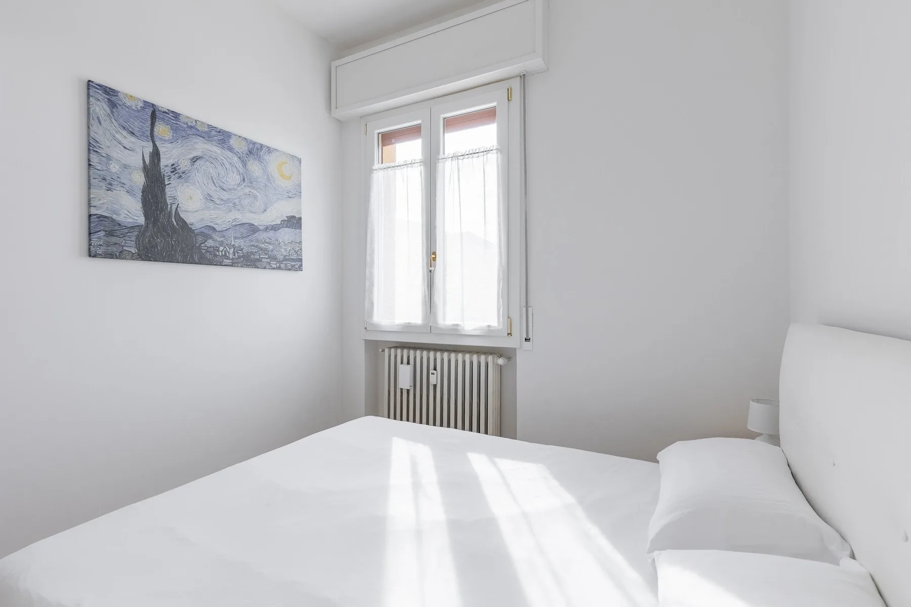 Un dormitorio luminoso con una cama bajo una ventana y un cuadro en la pared.